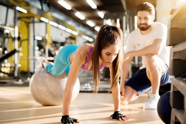 beautiful sporty woman doing push ups with fitness ball in gym, muscular personal trainer assisting her - Photo, Image