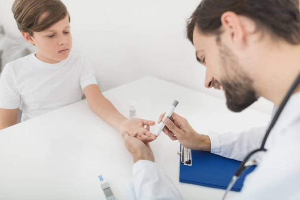 The doctor takes a blood sample from the boy to check it for sugar. The boy patiently endures the procedure - Fotoğraf, Görsel