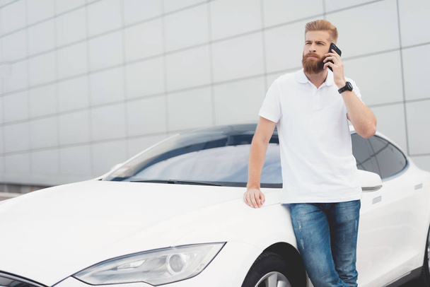 A young guy with a beard talking on the phone near his electric car. - Foto, Imagem