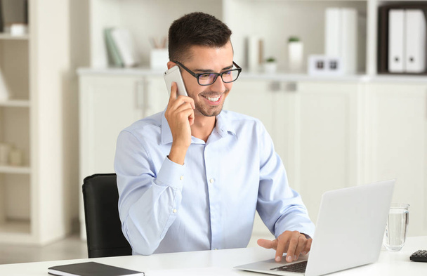 Attractive man with laptop talking on phone in office - Foto, imagen