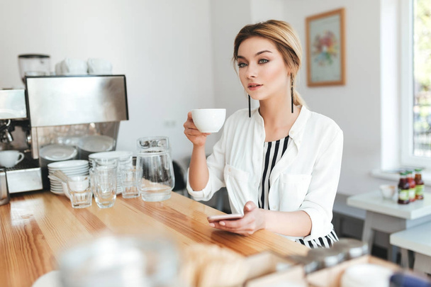 Porträt eines jungen Mädchens, das nachdenklich mit einer Tasse Kaffee und einem Handy in der Hand im Café zur Seite blickt. schöne Dame mit blonden Haaren sitzt am Tresen und trinkt Kaffee im Café  - Foto, Bild