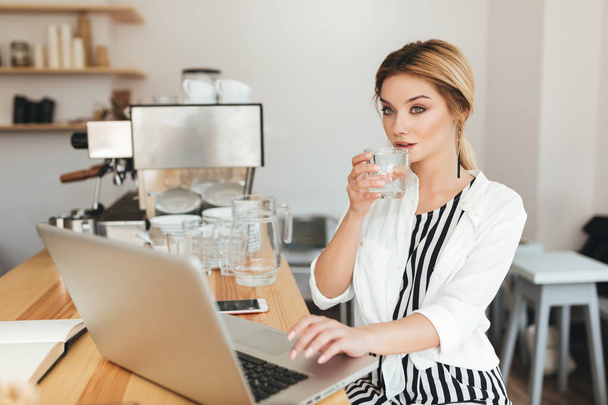 Nice girl sitting at the counter with glass of water in hand and thoughtfully looking aside in restaurant. Portrait of beautiful lady with blond hair working on her laptop in coffee shop - Foto, Bild