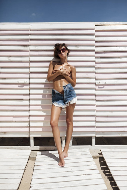 Portret pięknej dziewczyny w Dżinsowe szorty i okulary przeciwsłoneczne, spędzać czas na plaży. Młoda Ładna dziewczyna stojąc, obejmujące jej piersi z rękami na plaży  - Zdjęcie, obraz