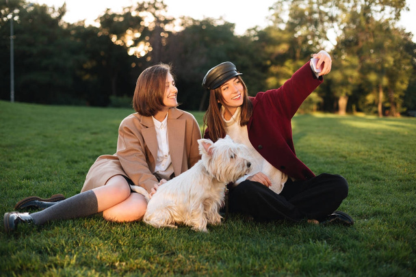 Portrait de deux belles filles assises sur l'herbe et prenant selfie sur téléphone portable tout en passant du temps avec leur chien mignon dans le parc
 - Photo, image