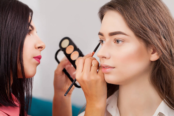 Kunstenaar doen professionele make-up van jonge vrouw in de buurt van de spiegel in schoonheid studio make-up - Foto, afbeelding