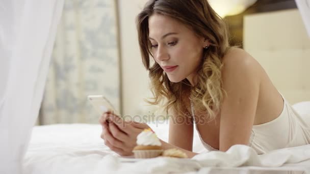 Woman lying on bed near cupcake and watching video on smartphone - Filmati, video