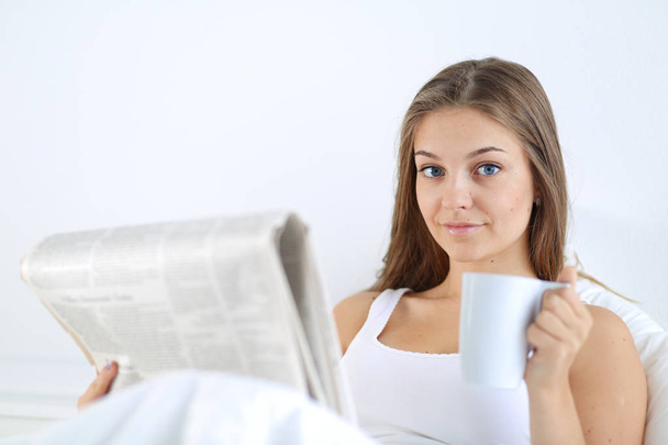 A pretty young woman reading the newspaper in bed and enjoying a cup of tea. - Foto, Imagen