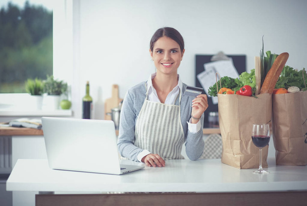 Smiling woman online shopping using computer and credit card in kitchen - Photo, Image