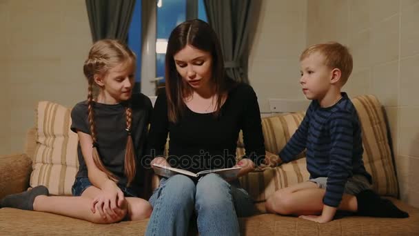 Mutter sitzt mit Tochter und kleinem Sohn auf der Couch und liest ihnen eine Geschichte vor, während sie auf der Couch im Wohnzimmer sitzen. - Filmmaterial, Video