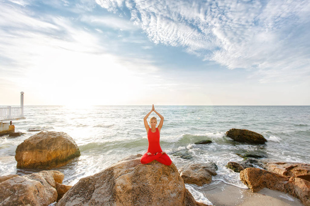 Žena v červeném obleku, cvičení jógy v kamenné při východu slunce u moře - Fotografie, Obrázek