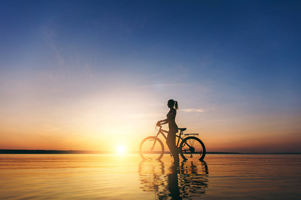 Silhouette d'une fille sportive en costume assis sur un vélo dans l'eau au coucher du soleil par une chaude journée d'été. Concept de fitness. Fond du ciel
 - Photo, image