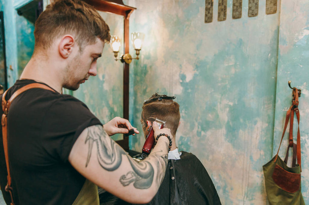 Close up shot of man getting trendy haircut at barber shop. Male hairstylist in tattoos serving client. - Foto, imagen