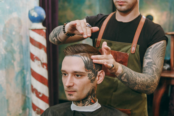 Close up shot of man getting trendy haircut at barber shop. Male hairstylist in tattoos serving client. - Zdjęcie, obraz