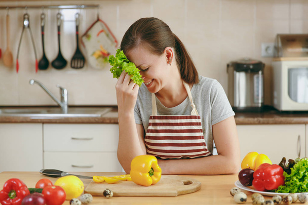 Una joven atractiva mujer en un delantal con hojas de lechuga riendo en la cocina. Concepto de dieta. Estilo de vida saludable. Cocinar en casa. Preparar alimentos
. - Foto, imagen