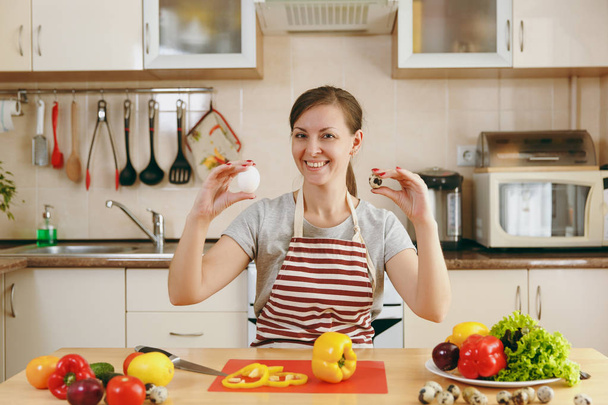 Een jonge gelukkig lachende vrouw in een schort kiest tussen kip en kwartel eieren in de keuken. Dieet concept. Gezonde levensstijl. Thuis koken. Eten bereiden. - Foto, afbeelding
