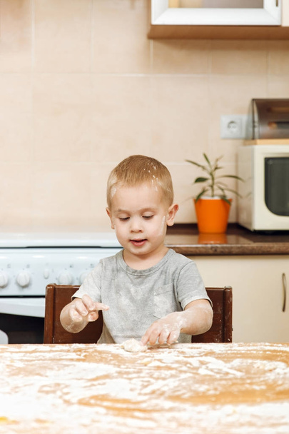 Μικρό παιδί αγόρι βοηθάει τη μητέρα να μαγειρεύουν Χριστούγεννα τζίντζερ μπισκότο στην ελαφριά κουζίνα. Ευτυχισμένο παιδί ξανθό σε γκρι T-shirt 2-3 χρόνια στο τραπέζι με ζύμη και το αλεύρι στο πρωί Σαββατοκύριακο στο σπίτι. - Φωτογραφία, εικόνα