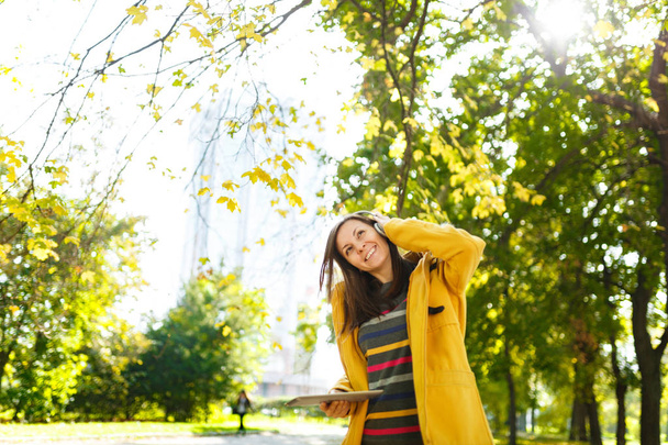 Güzel bir mutlu neşeli kahverengi saçlı kadın sarı ceket ve çizgili longsleeve bir tablet ile sıcak bir günde ellerini ve beyaz kulaklık sonbahar Şehir Parkı içinde sevinir. Altın yaprak. - Fotoğraf, Görsel