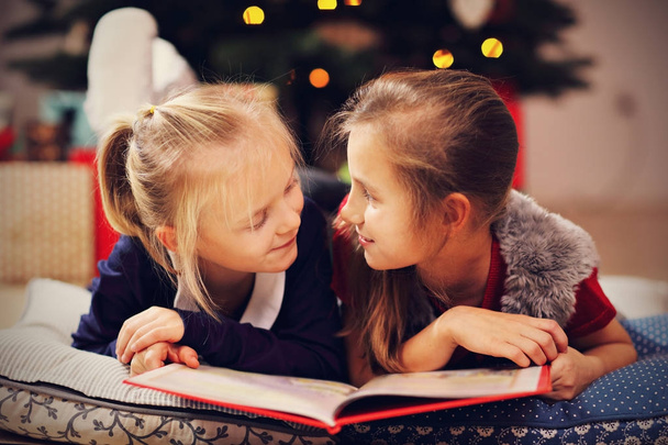 Two cute little sisters reading story book together under Christmas tree - Foto, imagen