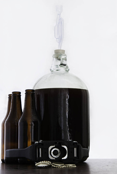 Fermented beer in a demijohn with airlock and brown bottles and over light background - Фото, изображение
