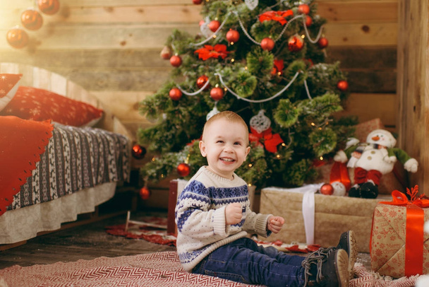 Gelukkig schattige kleine babyjongen gekleed in trui en jeans versieren kerstboom met speelgoed in houten kamer thuis. Kind met een goed humeur. Nieuwjaar. Lifestyle, familie en vakantie 2018 concept - Foto, afbeelding