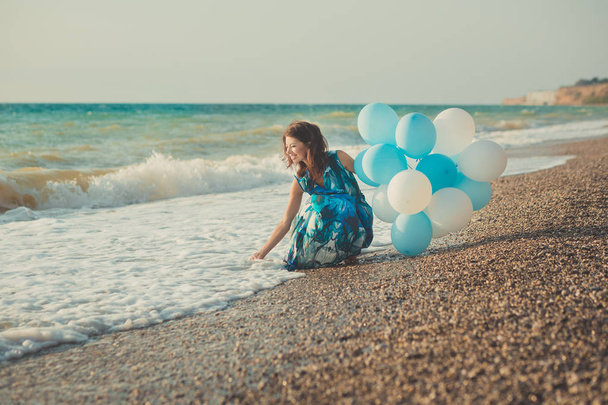 Amazing sensual lady woman with brunette hairs and ice blue eyes with white balloons posing sit for camera on summer sandy beach wearing fancy modish colourful dress barefoot - Photo, Image