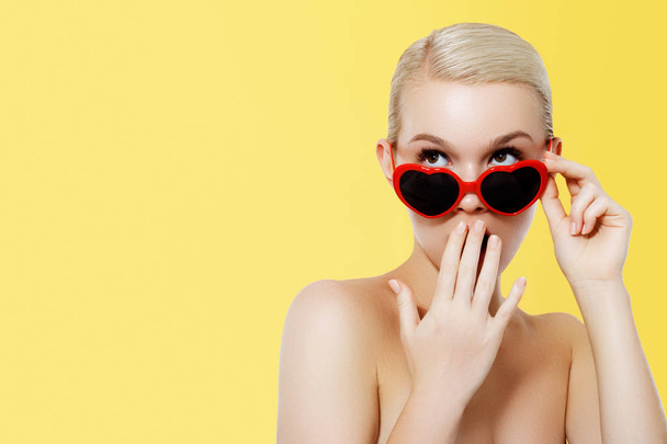 Valentine's Day concept. Fashion Model girl isolated over yellow background. Beauty stylish blonde woman posing in heart shaped sunglasses. Casual style with beauty accessories. High fashion style - Photo, Image