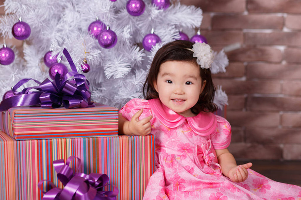 Asian pin up little cute girl wearing pink casual dress posing close to new year christmas white tree with violet purple balls toys and colourful presents - Zdjęcie, obraz