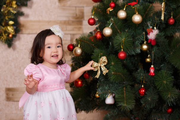 Asian little cute girl wearing casual dress posing close to new year christmas green classic tree with balls toys and colourful presents.Happy holidays with santa claus and kid child - Zdjęcie, obraz