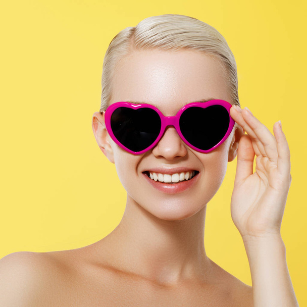 Valentine's Day concept. 14 february. Fashion Model girl isolated over yellow background. Beauty stylish blonde woman posing in heart shaped sunglasses. Casual style with beauty accessories - Fotoğraf, Görsel