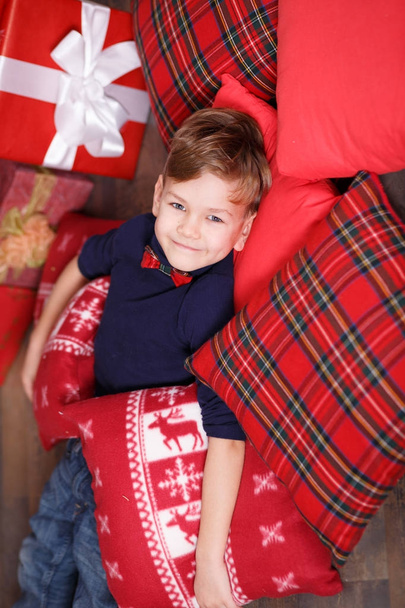 Handsome cute boy celebrating New Year Christmas alone close to xmas tree on red pillow posing in studio decoration wearing jeans and blue shirt with jazzbow tie - Фото, зображення