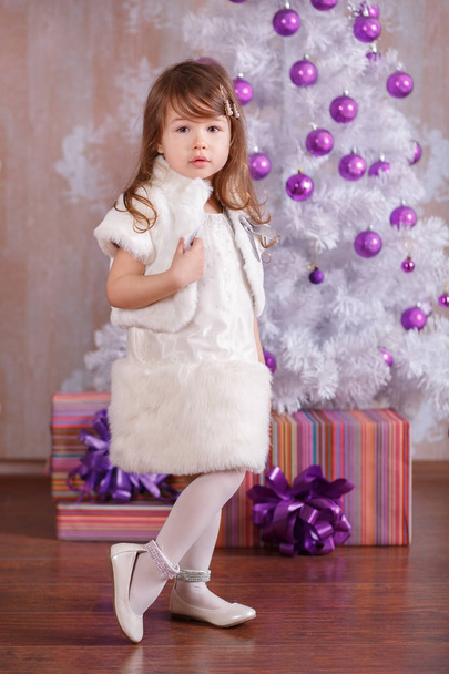 Young brunette dolly lady girl stylish dressed in cosy evening official white dress fur jacket strap shoes smiling posing in studio winter christmas tree and presents with pout lips and pink cheeks. - Foto, afbeelding