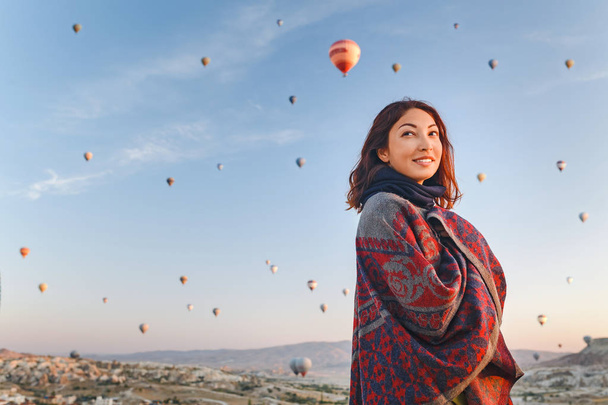 Kobieta w tradycyjnym Ponczo ubrania oglądać wspaniały widok, kolorowe balony latające nad doliną w regionie Kapadocja, Turcja. - Zdjęcie, obraz