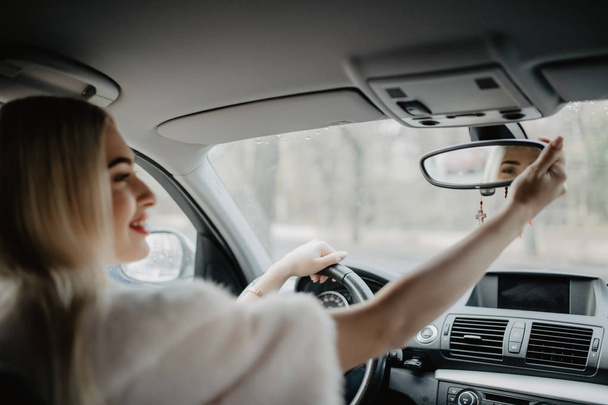 Όμορφη νεαρή γυναίκα ψάχνει στον καθρέφτη κατά την οδήγηση αυτοκινήτου στο δρόμο - Φωτογραφία, εικόνα