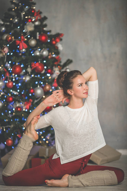 Beautiful flexible young woman is doing stretching exercise near decorated fashion xmas tree at home, sports and holiday concept - Foto, Imagen