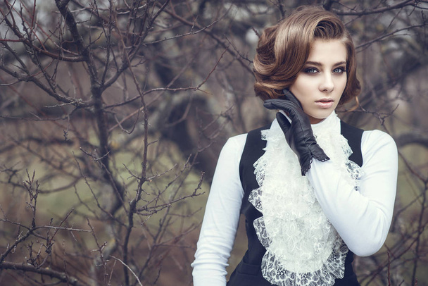Portrait of gorgeous young woman with elegant Victorian hairstyle and beautiful make up wearing old-fashioned gown with jabot standing at the dry tree holding her hand in leather glove at her cheek - Photo, Image