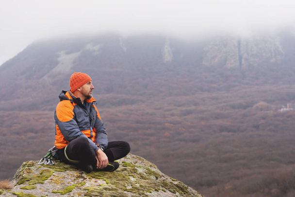 Hipster - ορειβάτης στο κάτω σακάκι και ένα πλεκτό κάλυμμα κάθεται και ακουμπά στην κορυφή ενός βράχου - Φωτογραφία, εικόνα