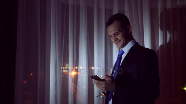 A young man in a business suit drinks alcohol, and makes selfie on his smartphone, stands at the window of the house at night. Window panorama, view of the city, night lights. 4k - Materiał filmowy, wideo
