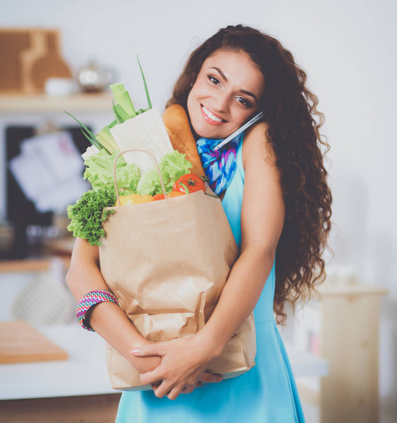 野菜と食料品の買い物袋を持っている若い女性キッチンに立つ - 写真・画像
