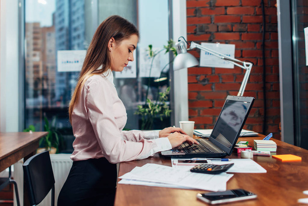 Nuori nainen yllään muodollinen vaatteita työskentelee kannettavan tietokoneen kirjoittamalla sähköposteja istuu hänen työpaikallaan
 - Valokuva, kuva
