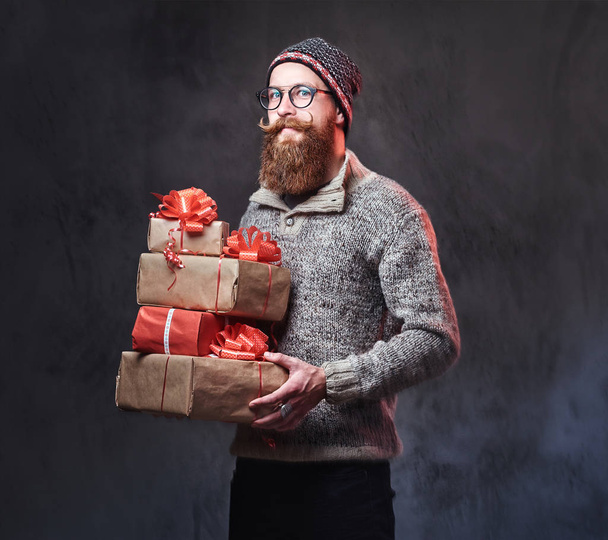 Retrato de macho barbudo em óculos vestidos com uma camisola de lã quente e um chapéu prende presentes de Natal sobre fundo cinza
. - Foto, Imagem