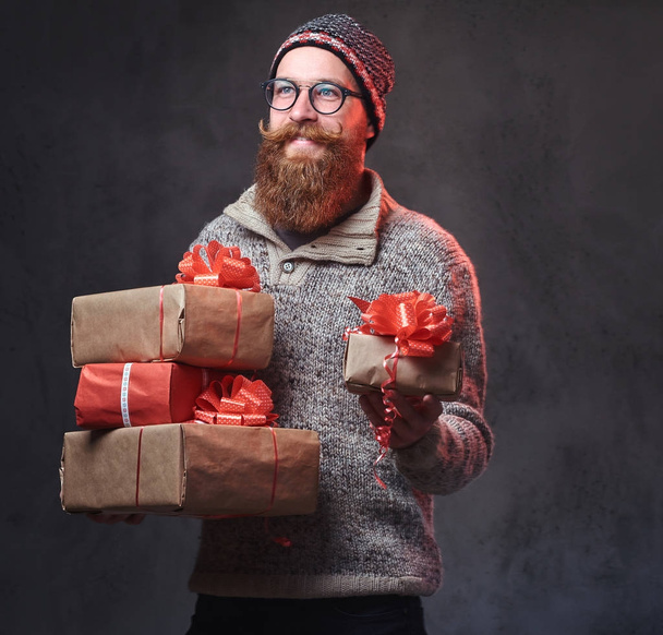 Portrait of bearded male in eyeglasses dressed in a warm wool sweater and a hat holds Christmas gifts over grey background. - Fotó, kép