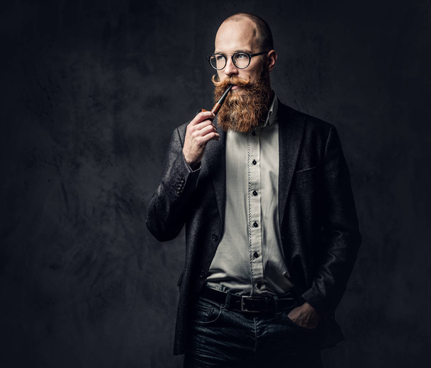 Redhead bearded male dressed in a suit and eyeglasses smoking tradition pipe over dark grey background. - Photo, Image