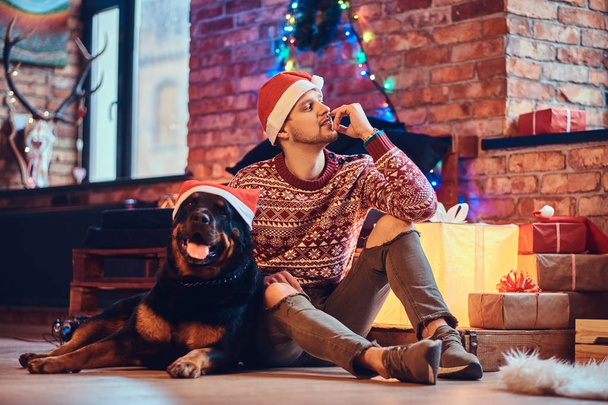 Atractivo macho hipster barbudo se sienta en el suelo con su perro Rottweiler en una habitación con decoración navideña
. - Foto, Imagen