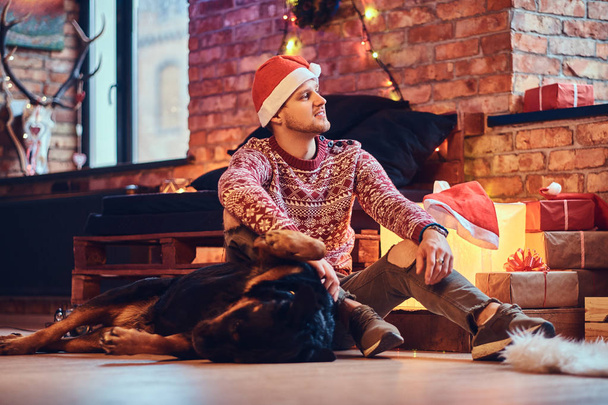 Houkutteleva parrakas hipsteri uros istuu lattialla rottweiler-koiransa kanssa huoneessa, jossa on joulukoriste.
. - Valokuva, kuva