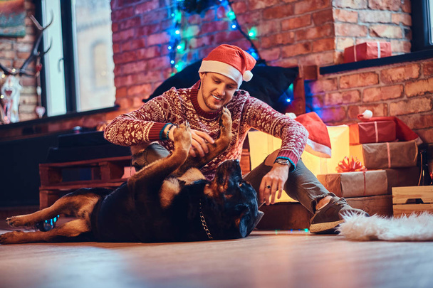 Atractivo macho hipster barbudo se sienta en el suelo con su perro Rottweiler en una habitación con decoración navideña
. - Foto, imagen