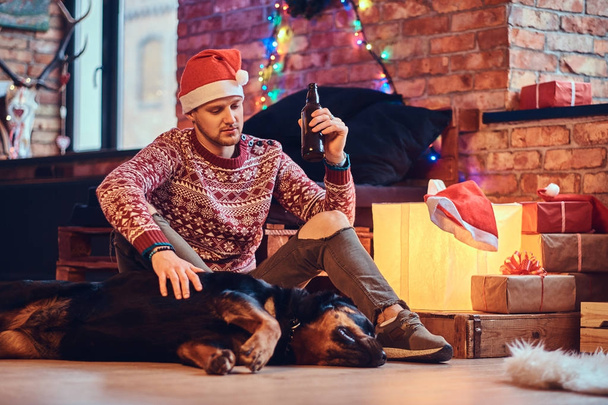 attraktiver bärtiger Hipster-Rüde sitzt mit seinem Rottweiler Hund auf dem Fußboden in einem Raum mit Weihnachtsdekoration. - Foto, Bild