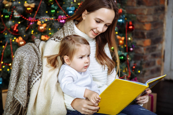 Pretty young mom reading a book to her cute daughter near Christmas tree indoors. - 写真・画像