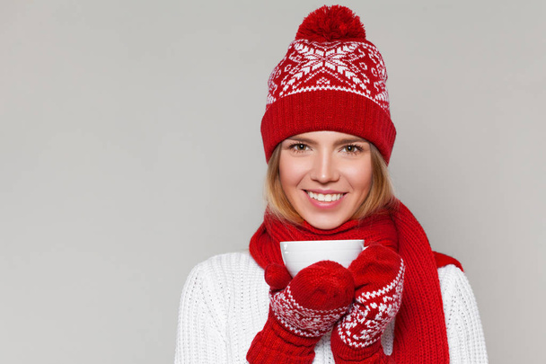 Ragazza invernale sorridente in cappello caldo lavorato a maglia e guanti che tengono una tazza in mano. Felice Natale donna, isolato su sfondo grigio
 - Foto, immagini