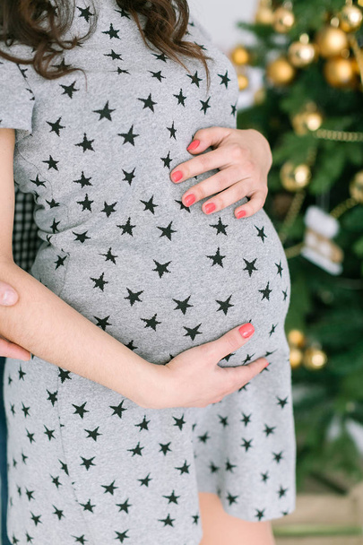 pregnancy, femininity, birth of children concept - closeup round belly of standing young future mother, hands of expectant lady gently support stomach, grey dress with stars pattern, selective focus - Photo, image