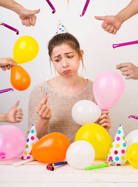 emotions, sentiments, birthday party concept. very sad disheveled girl, that is sulking, she is surrounded by colorful balloons, party hats with dots, hands with thumbs up and whistles - Foto, Imagem